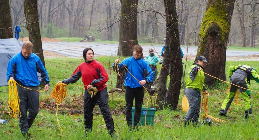 A group of people stand in a green wooded area and practice skills with ropes. 